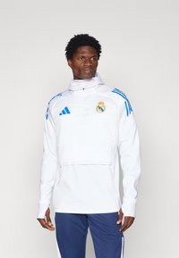 Witte trui met blauwe accenten. Heeft een hoge kraag, ritsluiting en geborduurd logo. Lichtgewicht, sportief ontwerp. Lange mouwen.