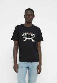 Crna majica kratkih rukava s grafikonom "Jack & Jones ARCHIVE" u bijeloj i zlatnoj boji. Majica je s okruglim izrezom i izrađena od mekanog pamučnog platna. Kombinirana s svijetloplavim trapericama.