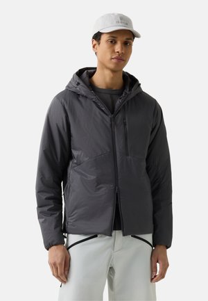 Mężczyzna ubrany w ciemnoszarą kurtkę z kapturem i białe spodnie z czarnymi zamkami błyskawicznymi, oraz jasnoszara czapka z daszkiem, stojący na neutralnym tle.