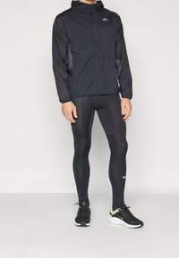 Czarna, lekka kurtka z zamkiem i kapturem, zestawiona z dopasowanymi czarnymi legginsami oraz czarnymi butami do biegania z żółtymi akcentami.