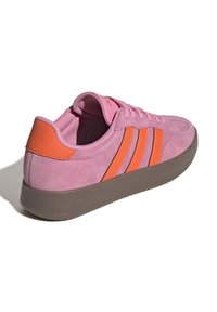 Rosa mockaskinns Adidas-sneakers med orangea ränder och hälflik, grå sula, sedd från bakre högra sidan på vit bakgrund.