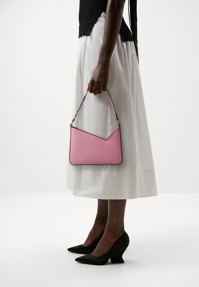 Femme tenant un petit sac à main rose avec une bandoulière en chaîne, portant une jupe midi blanche, un haut noir et des talons noirs pointus à talons blocs.