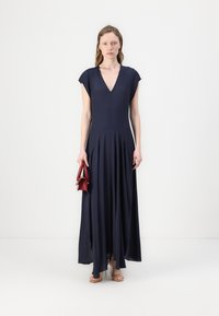 Námořnickě modré maxi šaty s výstřihem do V, krátkými rukávy a volnou siluetou. Model drží malou červenou kabelku. Hladký materiál s jemným drapováním.