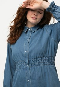 Camicia in denim con colletto, vita elasticizzata e maniche lunghe. Presenta bottoni e un colore azzurro chiaro. Texture liscia.