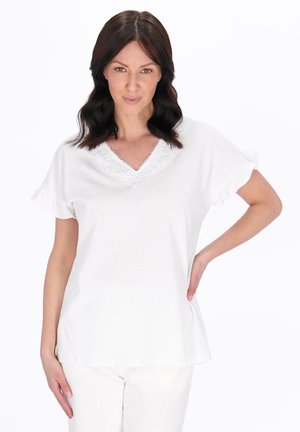 Femeie cu păr închis la culoare, lung până la umeri, purtând un top alb cu decolteu în V și tiv din dantelă și pantaloni albi, pozând cu o mână pe șold pe fundal alb.