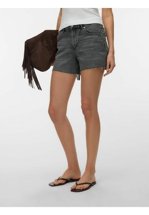 Fekete farmer rövidnadrágot, fehér felsőt és fekete papucsot viselő nő, barna rojtos táskát tart egy egyszínű fehér háttér előtt.