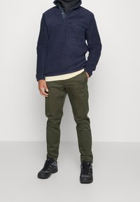 Forro polar azul marino con media cremallera y bolsillo; combinado con pantalones ajustados de color verde oliva y zapatos de senderismo negros con detalles grises.