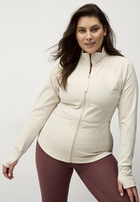 Beige ritsjack met hoge kraag, lange mouwen en duimgaten, gecombineerd met bordeaux leggings. Glad, rekbaar stoffenontwerp.