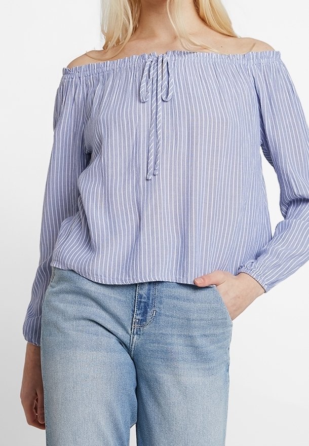 Femme portant un chemisier à rayures verticales bleu et blanc, épaules dénudées, manches longues, et un jean bleu clair, main dans la poche.