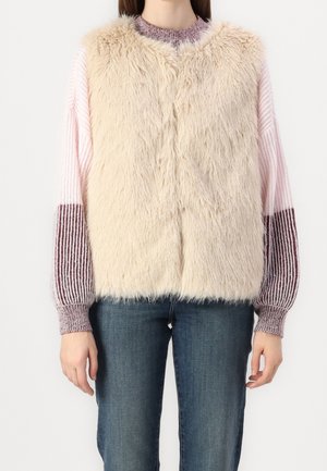 Vrouw draagt een beige mouwloze faux-fur gilet over een roze en bordeauxrood gestreepte gebreide trui met blauwe spijkerbroek, staand tegen een effen achtergrond.