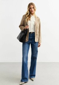 Beige suede blazer over a white blouse with a bow, paired with flared blue jeans and patterned pointed-toe heels. Black bag in hand.