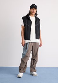 Sort ærmeløs jakke med hætte, grå accenter; hvid t-shirt under; brune bukser med blå sidelister; hvide sneakers.