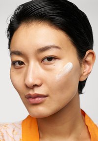 Hudvårdsprodukt applicerad på kinden, krämig vit textur på slät hud. Kort svart hår, bär en orange krage, neutral bakgrund.