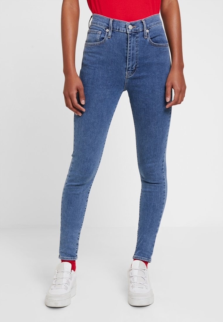 Jeans de mezclilla azul ajustados con cintura alta, que cuentan con cinco bolsillos, pasadores para cinturón y herrajes en tono plateado. Llevados con zapatillas blancas.