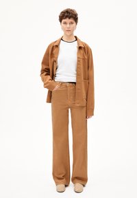 Veste en coton beige sur un haut blanc, associée à un pantalon large beige. Le tissu semble texturé, avec des détails de boutons et de poches.