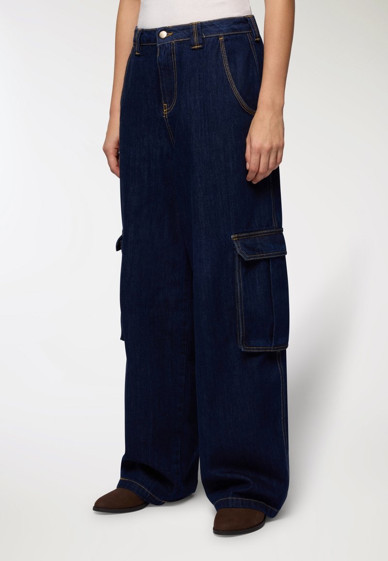 Jeans cargo a gamba larga in denim scuro con due tasche laterali, cuciture gialle a contrasto e vestibilità comoda. Indossate scarpe marroni basse.