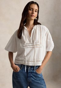 Γυναίκα με μακριά σκούρα μαλλιά που φοράει λευκή μπλούζα με δαντέλα και μπλε τζιν, στέκεται με τα χέρια στις τσέπες μπροστά σε ουδέτερο φόντο.