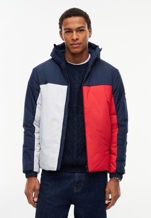Ung mand iført en hættetrøje med marineblå, hvid og rød farveblok, marineblå sweater under og mørkeblå jeans.