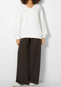 Felpa bianca a collo a V con maniche lunghe a palloncino, abbinata a pantaloni larghi marroni e calzature color crema. Materiale liscio e morbido.