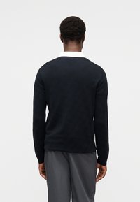 Μπλε σκούρο πλεκτό πουλόβερ με υφή, στρογγυλή λαιμόκοψη και μακριά μανίκια, σε συνδυασμό με γκρι παντελόνι. Μινιμαλιστικός σχεδιασμός χωρίς εμφανείς διακοσμητικές λεπτομέρειες.