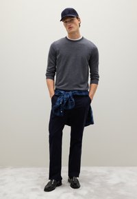 Maglione grigio con colletto bianco, camicia a quadri blu navy legata in vita, pantaloni scuri e scarpe nere. Texture liscia e design casual.