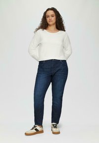 Biała bluzka z długim rękawem, ciemnoniebieskie dżinsy i kremowe sneakersy z brązowymi akcentami. Model stoi prosto na jasnym tle.