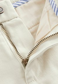 Tela de color crema con una cremallera visible y un detalle de cuello a rayas azules. La textura es suave con un ligero patrón acanalado.