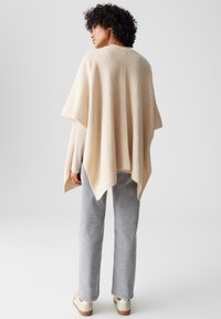 Beige gestrickter Poncho mit gerippter Textur, lockerer Form und subtilen Nahtdetails, kombiniert mit grauen Hosen und hellen Schuhen.