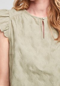Blouse vert clair en tissu texturé avec un motif de feuilles, dotée de manches courtes à volants et d'un décolleté en forme de goutte avec un bouton.