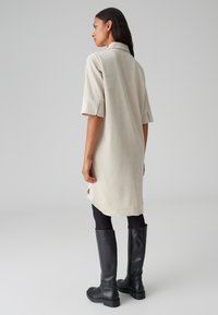 Abito camicia beige chiaro con colletto, maniche corte e spacchi laterali. Abbinato a stivali neri aderenti al ginocchio. Tessuto morbido.