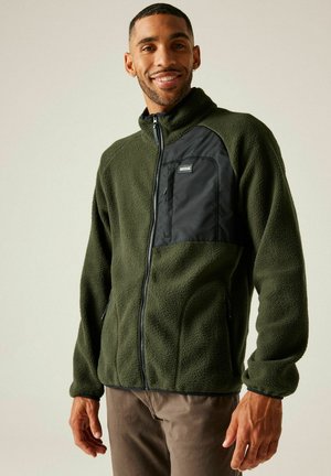 Man smiling while wearing an olive green fleece jacket with a black chest pocket and brown pants against a neutral background.