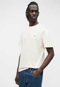 Μπεζ βαμβακερό t-shirt με κοντά μανίκια, που διαθέτει ένα μικρό πράσινο λογότυπο "II" στο στήθος. Συνδυασμένο με μπλε τζιν παντελόνι.