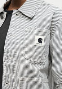 Striped grey and white jacket pocket with Carhartt logo patch on chest over a black shirt.