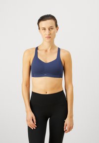 Sujetador deportivo azul marino con escote redondeado y tirantes anchos. Presenta una tela suave y un sutil detalle de logo. Combinado con leggings negros de cintura alta.