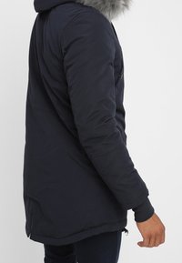 Person i en mörk marinblå vinterkappa med en huva kantad i grå päls, stående i profil med sin högra hand synlig.