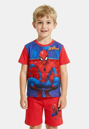 Jeune garçon souriant portant un t-shirt Spider-Man rouge et bleu ainsi que des shorts rouges avec un petit motif Spider-Man sur la jambe gauche.