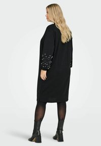 Robe pull noire à manches longues avec des accents de perles blanches sur les poignets. Coupée en dessous du genou avec des fentes sur les côtés. Portée avec des bottines noires.