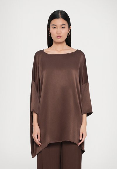 Haut oversized en satin marron avec un large décolleté et des manches trois-quarts, présentant une texture lisse et un design d'ourlet haut-bas.