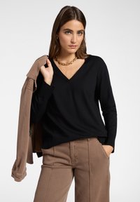 Frau in schwarzem V-Ausschnitt-Pullover und braunen Hosen hält eine passende braune Jacke über ihrer Schulter und trägt eine goldene Kette.