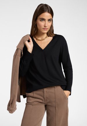 Una donna in un maglione nero a V e pantaloni marroni tiene una giacca coordinata marrone sulla spalla, indossando una collana di catena dorata.