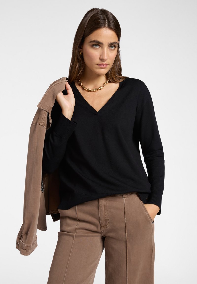 Frau in schwarzem V-Ausschnitt-Pullover und braunen Hosen hält eine passende braune Jacke über ihrer Schulter und trägt eine goldene Kette.