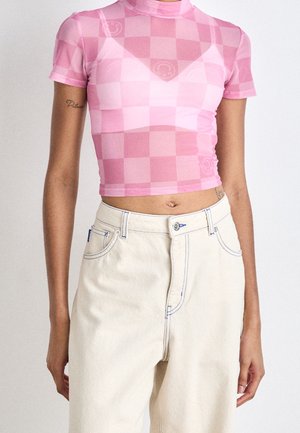Personne mince portant un crop top à manches courtes à carreaux roses et un jean taille haute écru avec des boutons et des poches visibles.