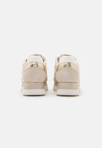 Ljust beige sneakers med mockatextur, gyllene metalliska detaljer på klackarna, vita gummisulor och rund tåsdesign. Utsikt bakifrån.