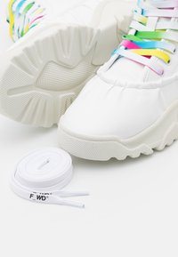 Baskets blanches avec semelles épaisses texturées et lacets arc-en-ciel pastel colorés, accompagnées d'une paire de lacets blancs roulés avec texte noir.