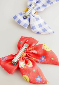 Två hårspännen: en blå och vit rutig rosett, en röd blommig rosett med leende ansikten, båda med metallclips på baksidan.