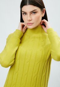 Maglione a collo alto lavorato a maglia di un brillante colore giallo, con un design a trecce, colletto e polsini a coste, e una vestibilità rilassata con fessure laterali.