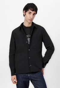 Svart knappedrakt skjorte med to brystlommer, lange ermer og klassisk krage, båret over en grafisk hettegenser, kombinert med mørke jeans.