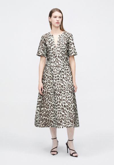 Γυναίκα ντυμένη με φόρεμα μήκους μέχρι το γόνατο με λεοπάρ τύπωμα και κοντά μανίκια, λευκές κάλτσες δαντέλα και μαύρα σανδάλια με ψηλό τακούνι, στέκεται σε λευκό φόντο.