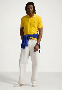 Άνδρας που φοράει κίτρινο πόλο μπλουζάκι, λευκά παντελόνια, λευκά παπούτσια, με μπλε πουλόβερ δεμένο στη μέση, στέκεται μπροστά σε ουδέτερο φόντο.