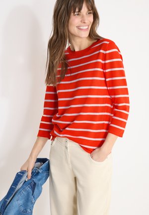 Smilende kvinde iført rød- og hvidstribet langærmet top og lysebeige bukser, som holder en blå denimjakke i venstre hånd.